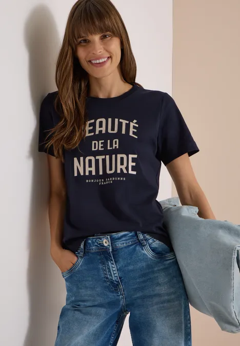 Eine lächelnde Frau trägt ein dunkelblaues Shirt mit Aufschrift „Beauté de la nature“, Jeans und hält eine Jacke.