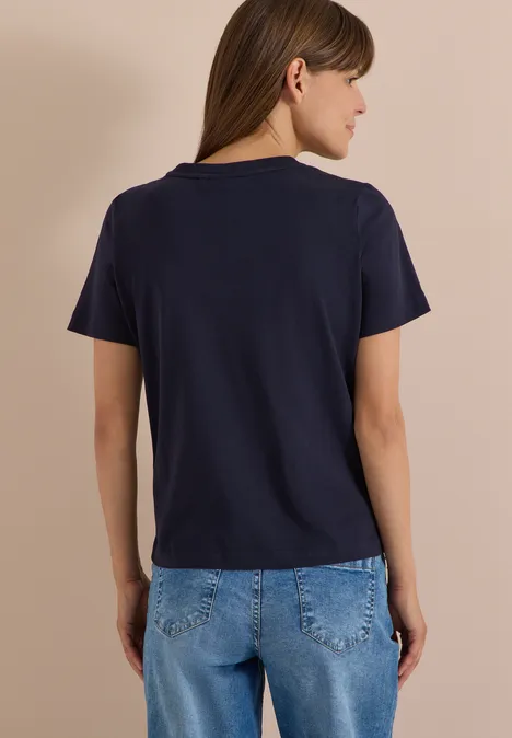 Person von hinten in dunkelblauem T-Shirt und Jeans vor beigem Hintergrund.