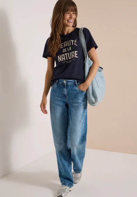 Eine lächelnde Frau mit dunklem T-Shirt und lockeren Jeans trägt eine Tasche über der Schulter.