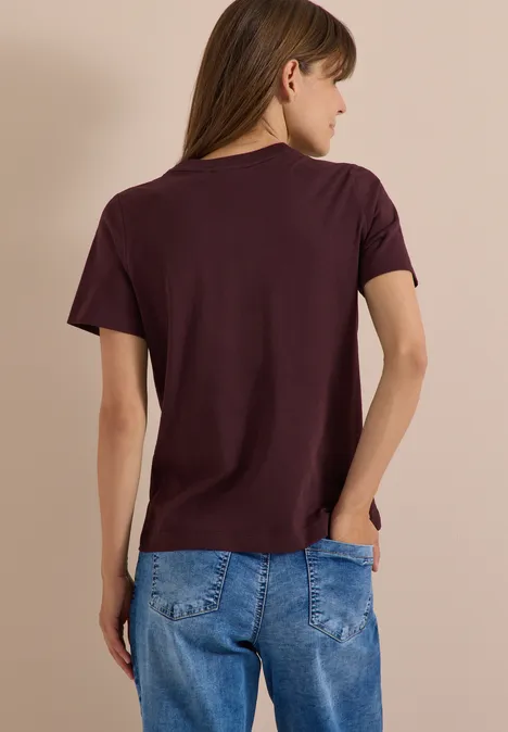 Eine Frau von hinten in weinrotem T-Shirt und blauen Jeans vor hellem Hintergrund.