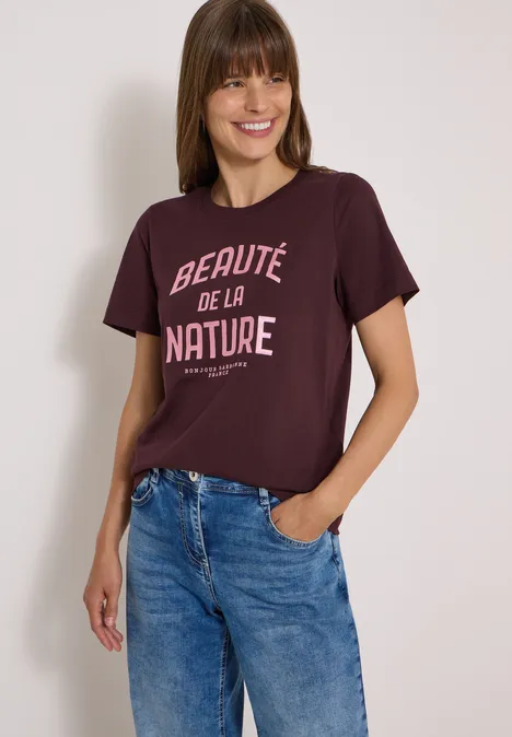 Frau in bordeauxrotem T-Shirt mit Aufschrift “Beauté de la nature” und lässigen blauen Jeans vor weißem Hintergrund.