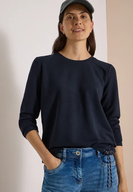 Eine Frau in blauer Mütze und Pullover mit Jeans, Hände in den Taschen.