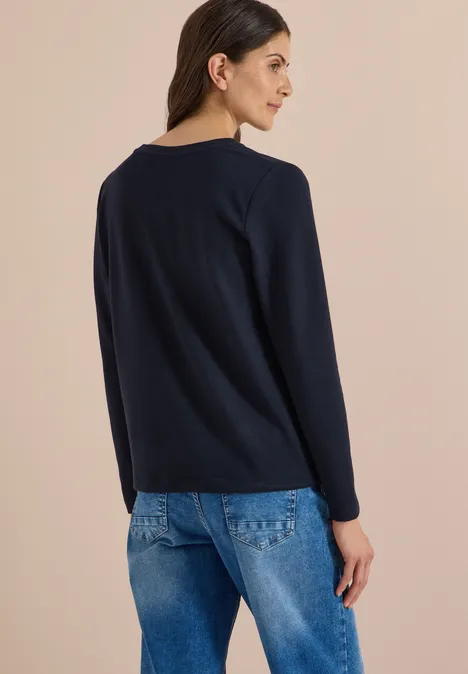 Frau in dunkelblauem Cardigan und Jeans, von hinten mit seitlicher Kopfwendung vor beigem Hintergrund.