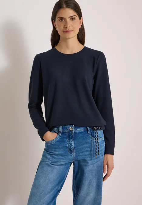 Frau in dunkelblauem Langarmshirt und blauen Jeans vor hellem Hintergrund.