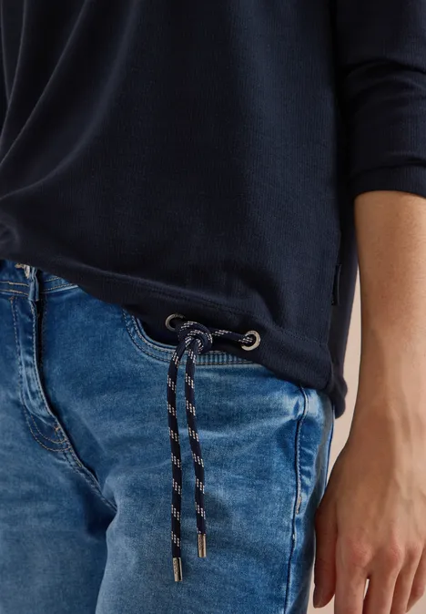 Person trägt dunkles Shirt und blaue Jeans mit geflochtenem Kordelzug an der Taille.