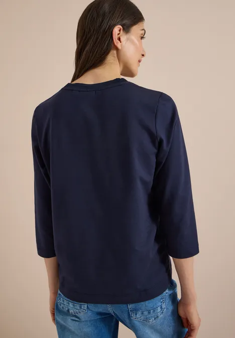 Rückenansicht einer Frau in navyblauem 3/4-Arm-Shirt und blauen Jeans vor beigem Hintergrund.