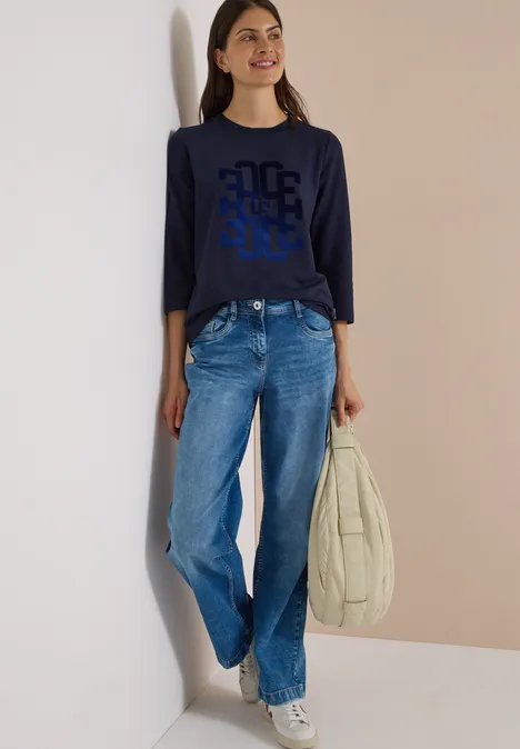 Frau lehnt an Wand, trägt marineblaues Langarmshirt mit Grafik, weite Jeans, weiße Sneaker und große Tasche.