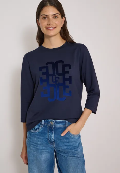 Eine lächelnde Frau in blauem Langarmshirt mit Grafik-Print und Jeans, eine Hand in der Hosentasche.