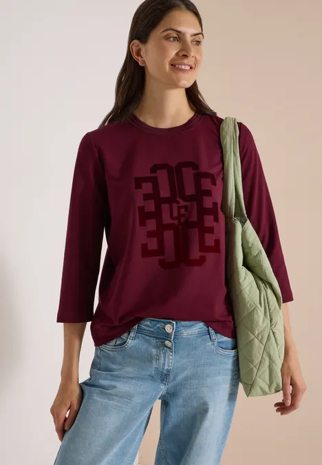 Frau mit burgunderfarbenem Shirt mit 3/4-Ärmeln, Jeans und grüner Stepp-Umhängetasche, lächelnd.