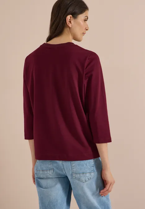 Frau von hinten mit dunkelrotem 3/4-Arm-Shirt und blauen Jeans vor beigem Hintergrund.