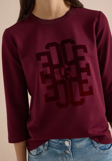 Frau trägt burgunderrotes Sweatshirt mit geometrischem Print und 3/4-Ärmeln, dazu blaue Jeans.