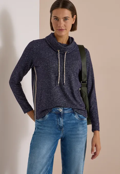 T-shirt souple à col et bande de renfort urban dark blue melange Frau in dunkelblauem Strickpullover mit Känguru-Kragen und Jeans, lässig vor beige Wand stehend.
