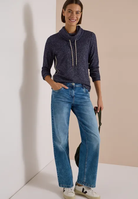 T-shirt souple à col et bande de renfort urban dark blue melange Frau in dunkelblauem Hoodie mit Kordeln, weiten Jeans und weißen Sneakers, lehnt an heller Wand.