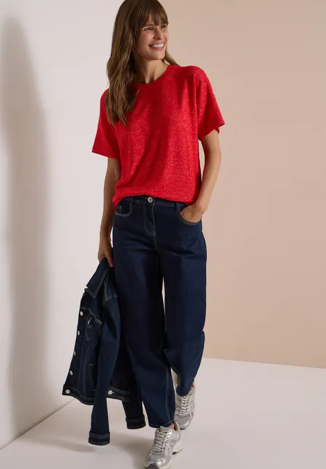 Shirt met verlaagde schouders, ronde hals en band flame red melange Frau in rotem T-Shirt, weiten Jeans und weißen Sneakers, hält eine Jeansjacke, steht vor hellem Hintergrund.