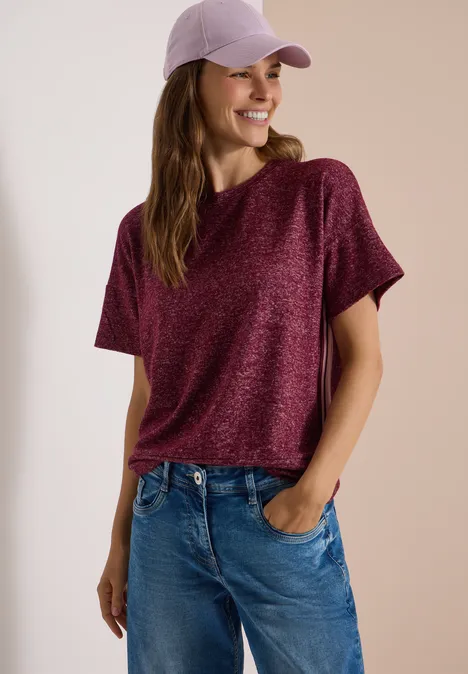Shirt met aflopende schouders, ronde hals en band red wine melange Frau trägt lila Cap, lockeres bordeauxfarbenes Shirt und Jeans, lächelt und hat Hände in den Taschen.