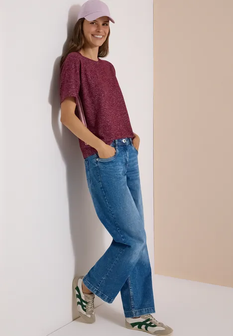 Shirt met aflopende schouders, ronde hals en band red wine melange Frau in lockerem Freizeitoutfit (lila Shirt, Jeans, Cap, weiße Sneaker) lehnt lächelnd an heller Wand.