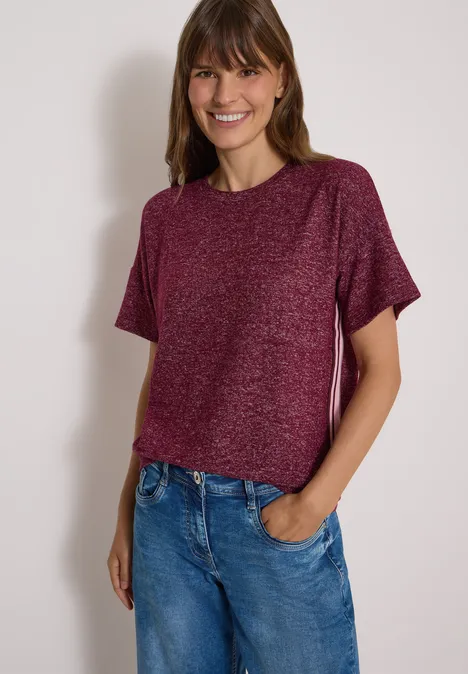 Shirt met aflopende schouders, ronde hals en band red wine melange Frau in rotem T-Shirt und blauer Jeans lächelt vor neutralem Hintergrund.