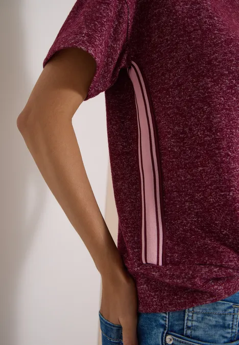 Shirt met aflopende schouders, ronde hals en band red wine melange Person trägt glänzendes bordeauxfarbenes Shirt mit weißen und rosa Seitenstreifen, Hand in Jeans-Tasche.