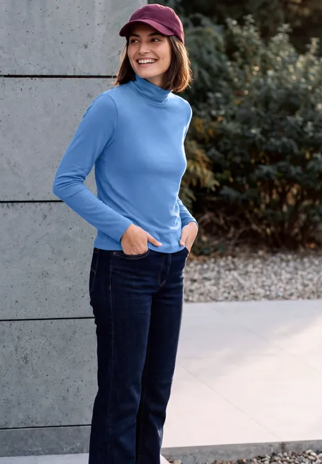 Femme souriante portant une casquette bordeaux, un pull à col roulé bleu et un jean, debout dehors, mains dans les poches.