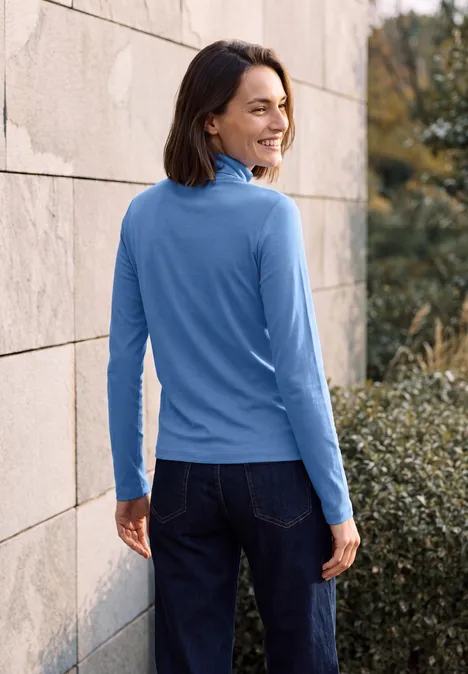 Femme en haut bleu et jean s'appuie sur un mur en pierre et sourit par-dessus son épaule.