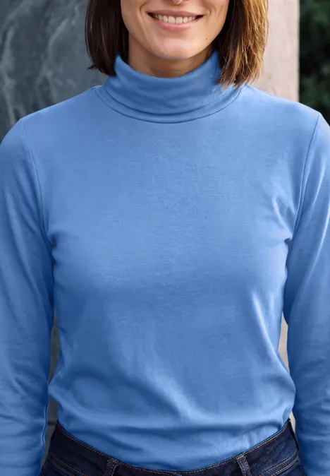 Une femme porte un col roulé bleu clair et un jean, souriante.