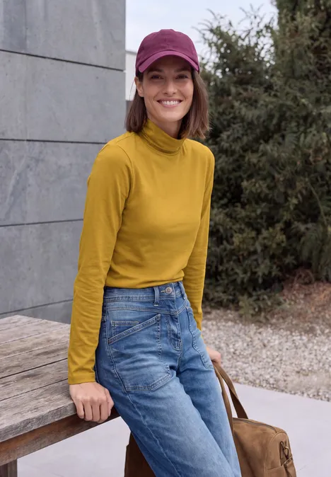 Langarm Shirt in Unifarbe mit Turtle Neck golden honey Eine Frau mit gelbem Rollkragenpullover, Jeans und pinker Mütze lehnt lächelnd an einer Holzbank.