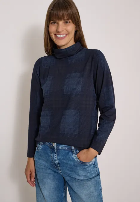 T-shirt à manches longues, col roulé et imprimé urban dark blue Femme souriante en pull à col roulé bleu foncé à carreaux et jean bleu, main dans la poche.