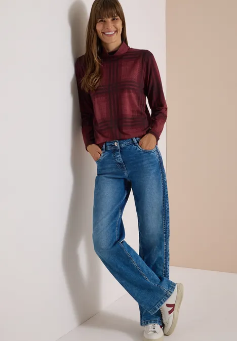 Eine lächelnde Frau lehnt an der Wand, sie trägt einen roten Karopullover, blaue Jeans und weiße Sneaker.