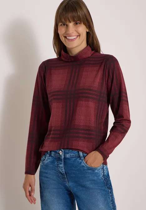 Eine Frau trägt ein bordeauxrotes Karopullover mit Rollkragen und blaue Jeans, lächelnd.