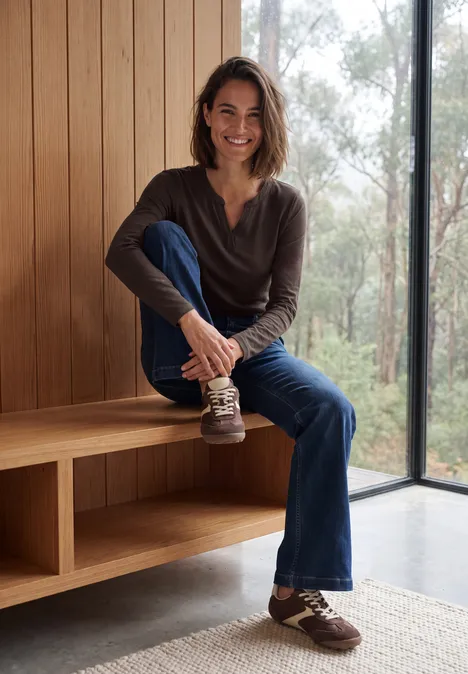 Shirt met split in de hals, in tuniekstijl truffle brown Vrouw in jeans en sneakers zit glimlachend op houten bank voor grote ramen met boszicht.