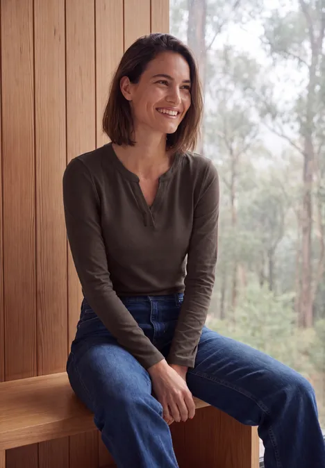 Shirt met split in de hals, in tuniekstijl truffle brown Vrouw zit glimlachend op een houten bank voor een raam met uitzicht op bebost landschap.