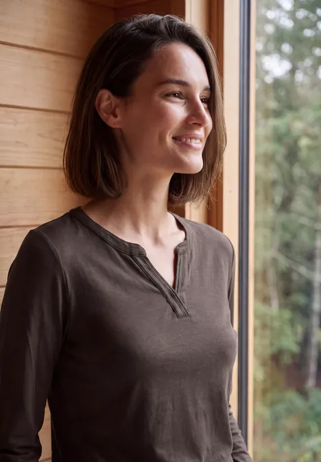 Shirt met split in de hals, in tuniekstijl truffle brown Een glimlachende vrouw staat bij het raam en kijkt uit op de groene tuin.