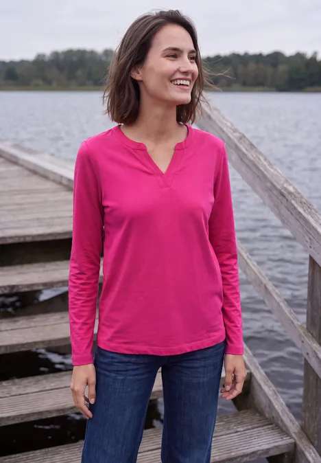 Shirt met split in de hals, in tuniekstijl pink flash Vrouw in roze shirt met lange mouwen en spijkerbroek lacht op een steiger bij het meer.