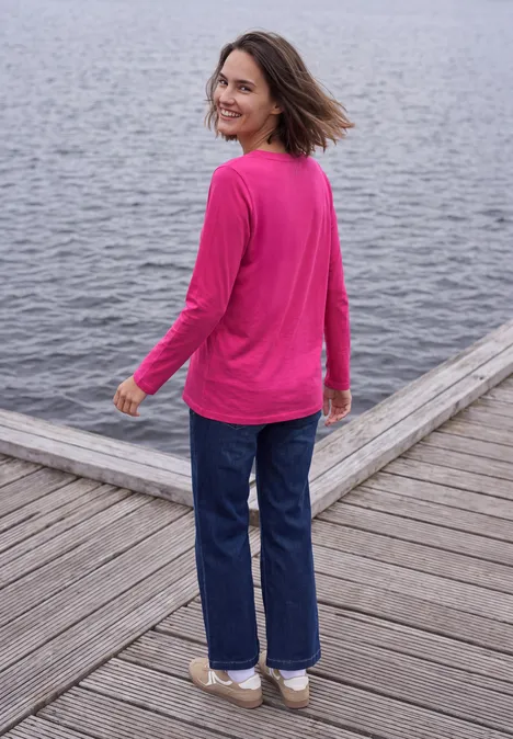 Shirt met split in de hals, in tuniekstijl pink flash Een vrouw in een roze trui en spijkerbroek lacht op een houten steiger aan het water.