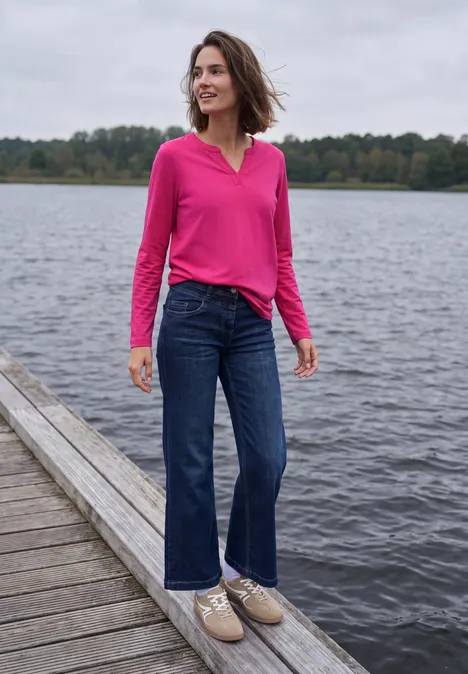 Shirt met split in de hals, in tuniekstijl pink flash Een vrouw in roze blouse en jeans staat lachend op een steiger bij een meer, bomen op de achtergrond.