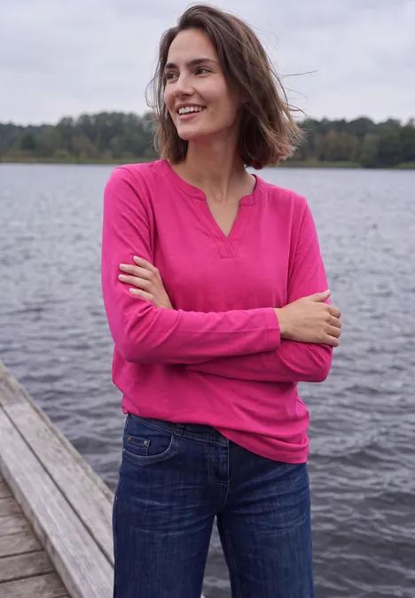 Shirt met split in de hals, in tuniekstijl pink flash Vrouw lacht op houten steiger bij meer, draagt roze shirt en spijkerbroek.