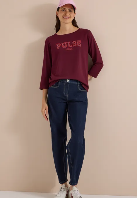 Frau mit rosa Cap, dunkelrotem Pulse-Shirt, blauen Jeans und weißen Sneakern vor hellem Hintergrund.