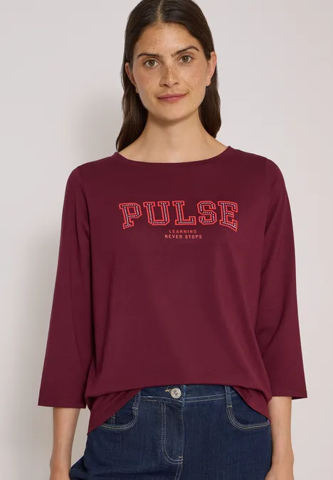 Frau in weinrotem Shirt mit 'PULSE'-Aufdruck und dunkler Jeans.