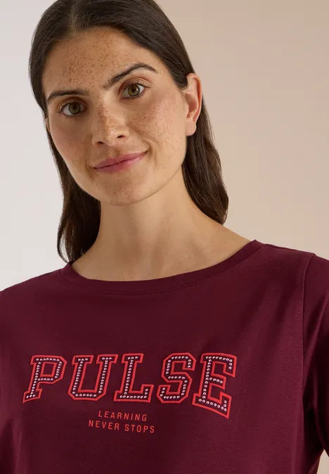 Frau mit dunklen Haaren trägt ein weinrotes Shirt mit weißer Aufschrift 'PULSE' und schaut freundlich.