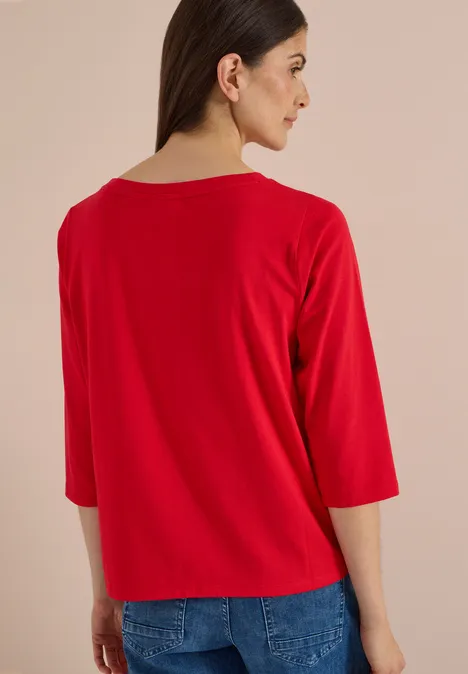 Frau in rotem Dreiviertelarm-Shirt und blauer Jeans, Rückenansicht, Blick seitlich.