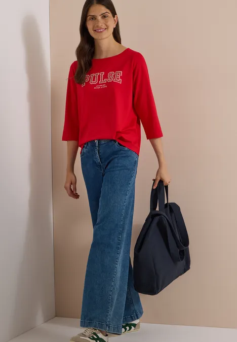 Junge Frau in rotem 3/4-Shirt mit 'POOLS'-Logo, blauen Schlagjeans, lächelnd mit dunkelblauer Tasche.