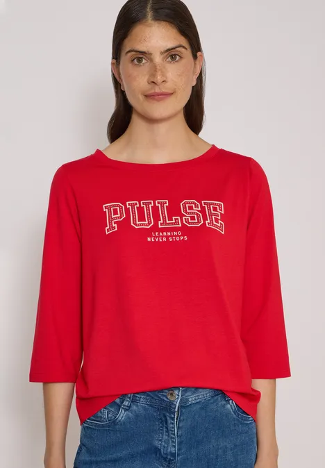 Junge Frau vor hellem Hintergrund trägt rotes Shirt mit 'PULSE'-Aufschrift und blaue Jeans.