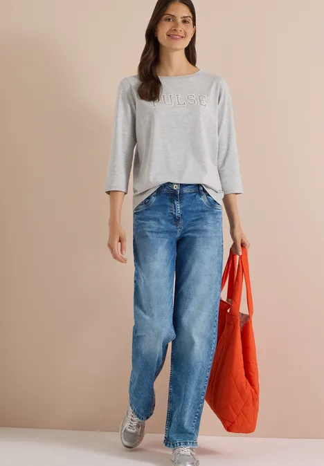Frau trägt graues 3/4-Shirt, blaue Jeans, graue Sneaker und orangefarbene Tasche