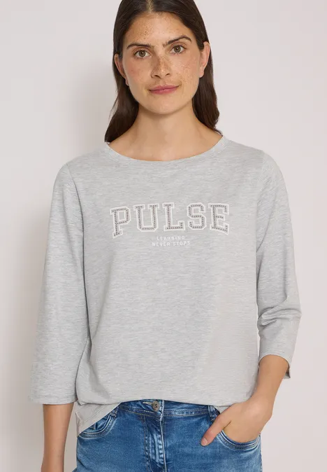 Junge Frau in grauem 'PULSE'-Shirt und Jeans, lächelnd vor hellem Hintergrund mit Hand in Tasche.
