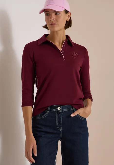 Frau in weinrotem Poloshirt mit Dreiviertelärmeln, blauen Jeans und rosa Baseballcap.