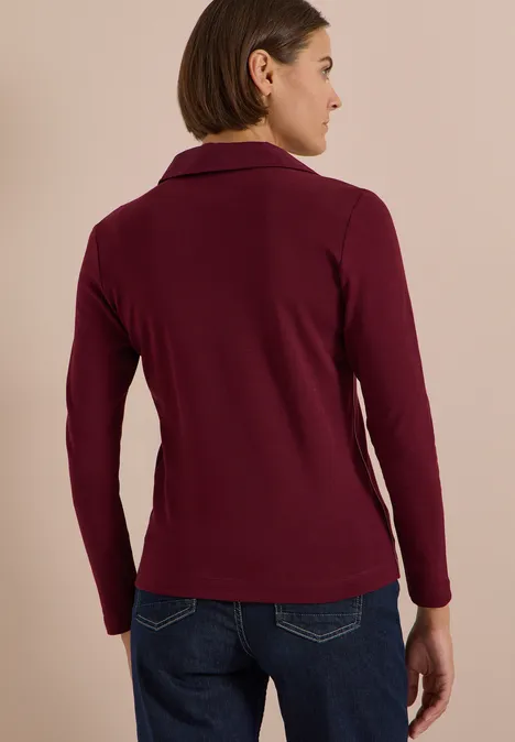 Frau von hinten in weinrotem Blazer und Jeans vor hellem Hintergrund.