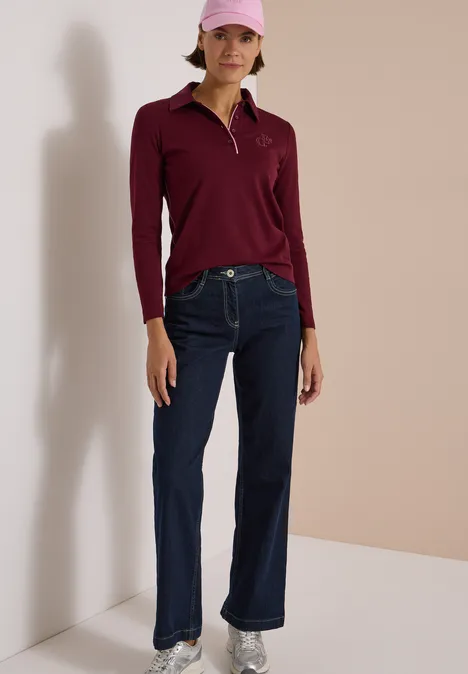 Frau trägt bordeauxfarbenes Polo, weite Jeans, rosa Cap vor neutralem Hintergrund.