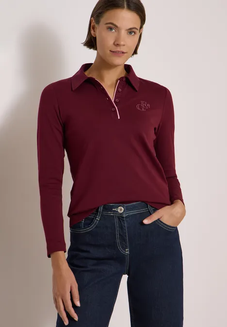 Frau in burgunderrotem Langarm-Poloshirt und blauer Jeans, locker stehend mit einer Hand in der Hosentasche.