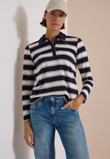 Frau trägt gestreiftes Langarm-Poloshirt, Jeans und Baseballcap, steht mit Hand in Tasche.
