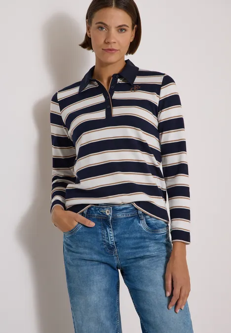 Frau trägt ein langärmeliges, gestreiftes Polo in Blau und Beige mit Jeans vor neutralem Hintergrund.